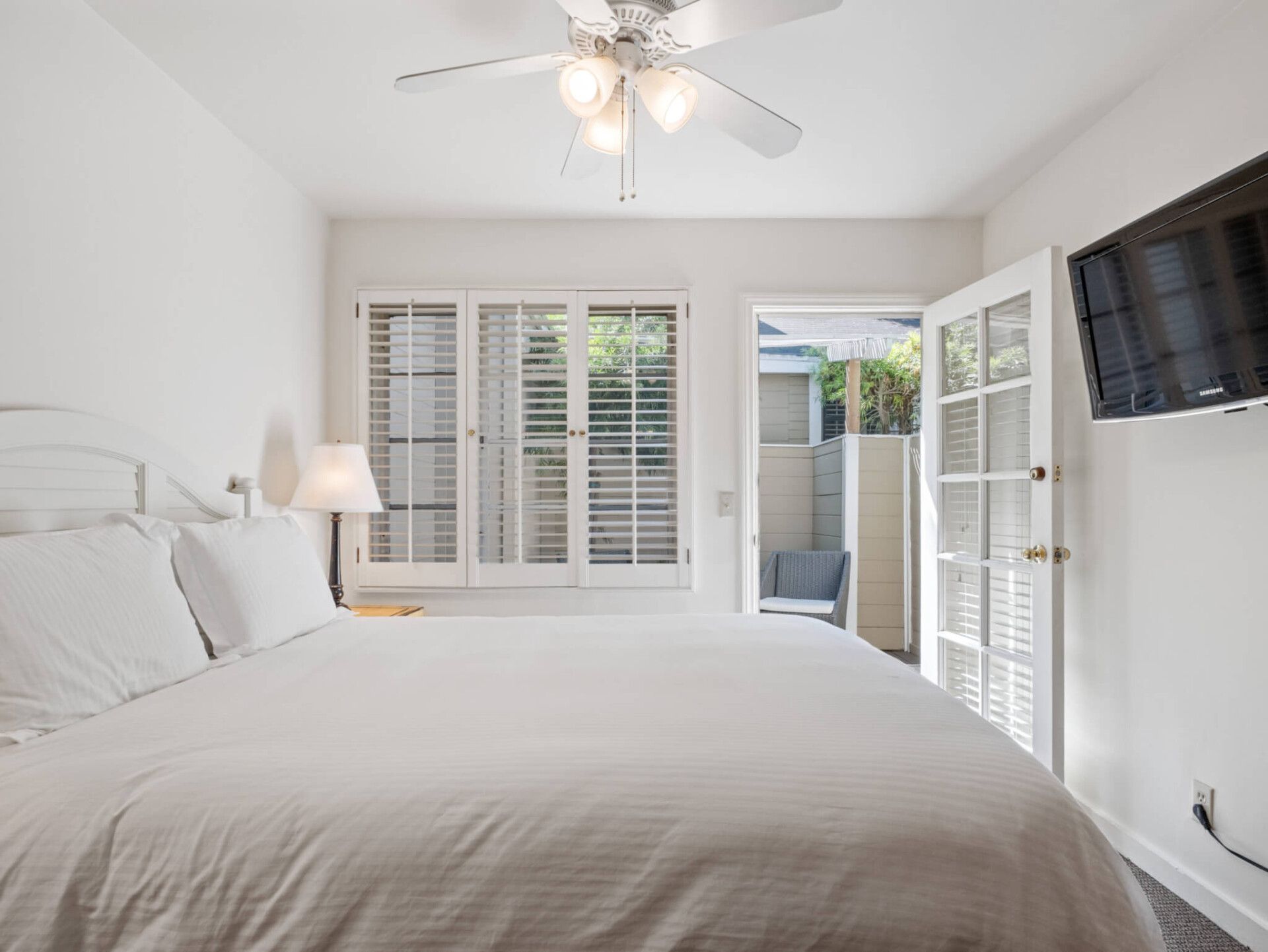 A bright bedroom with a white bed, ceiling fan, lamp, shuttered window, and a glass door leading to a patio with a chair.