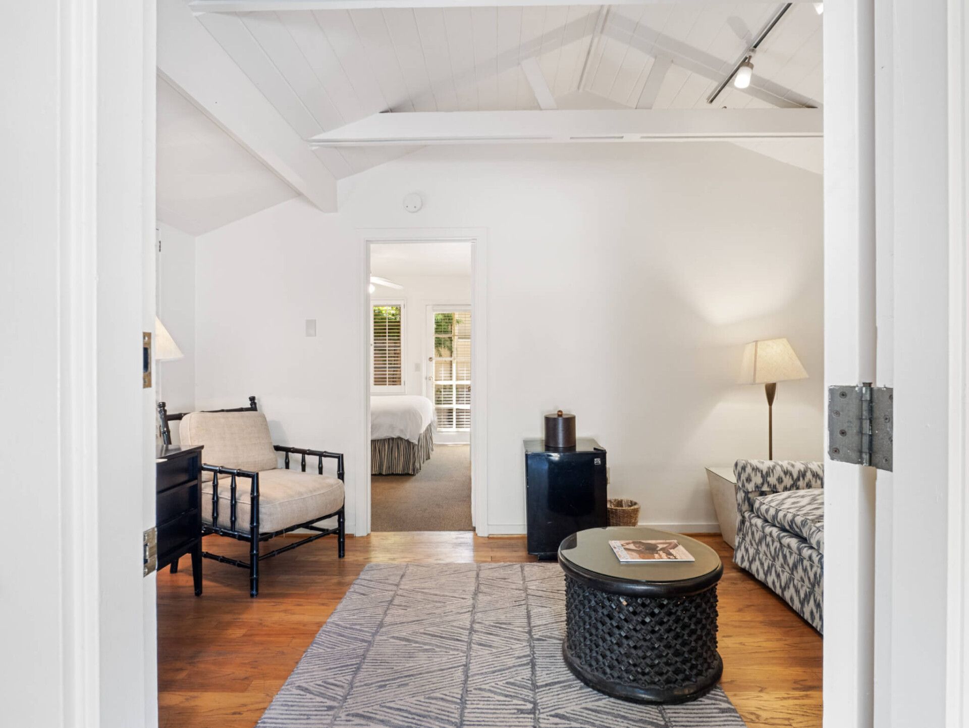 A bright, furnished room with a wooden floor, armchair, small round coffee table, and a doorway leading to another room.