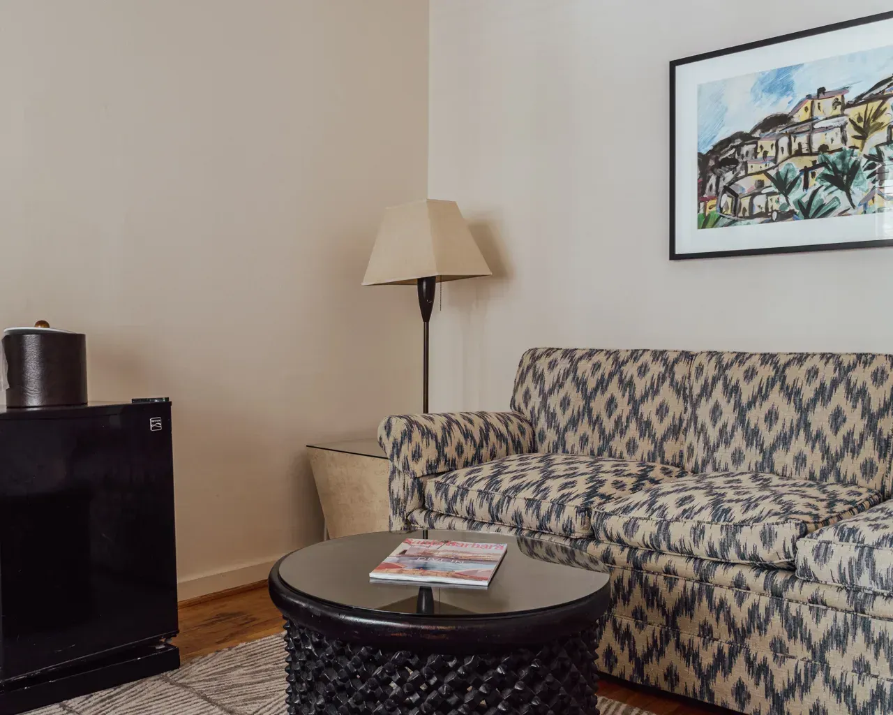 A patterned sofa with a round textured table in front, a lamp, and a black mini-fridge in a neutrally decorated room.