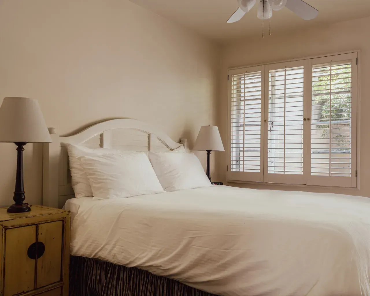 A tidy bedroom with a white bed, two bedside lamps, a nightstand, and window shutters on a neutral-colored wall.