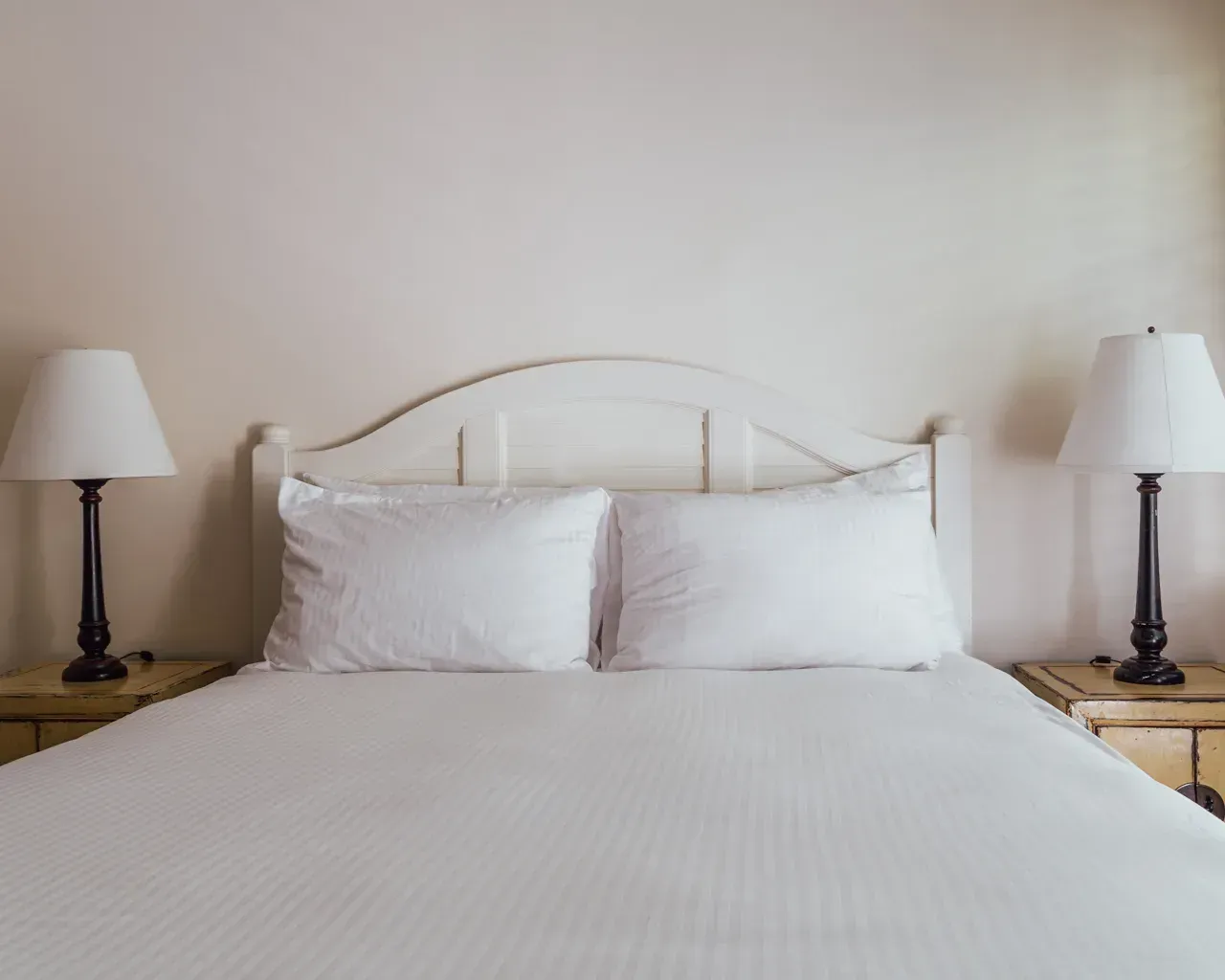A neatly made bed with white bedding, pillows, and a wooden headboard, flanked by two bedside lamps on matching tables.