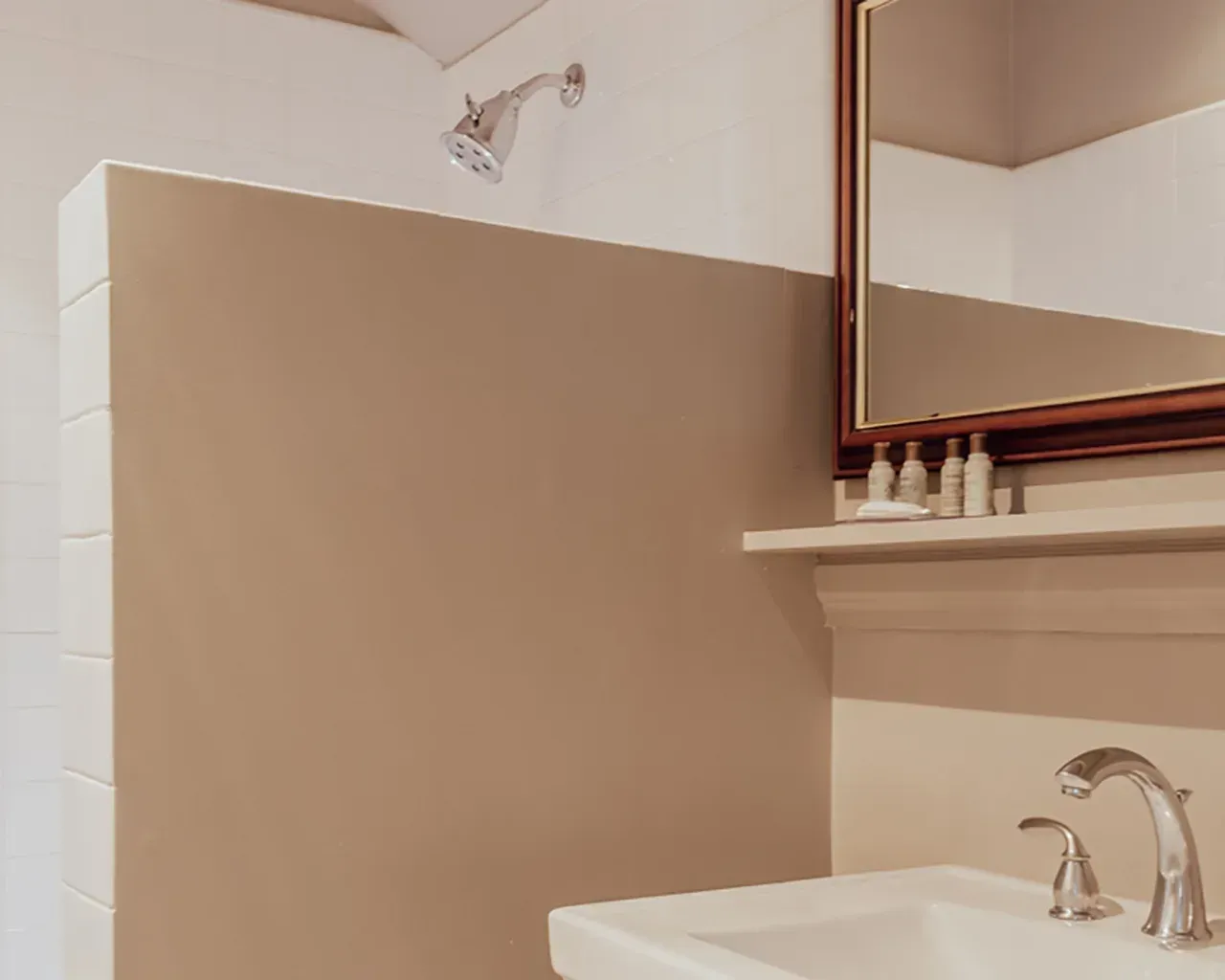 A modern bathroom vanity with a white sink, chrome faucet, small shelf with bottles, a mirror, and a beige shower wall.