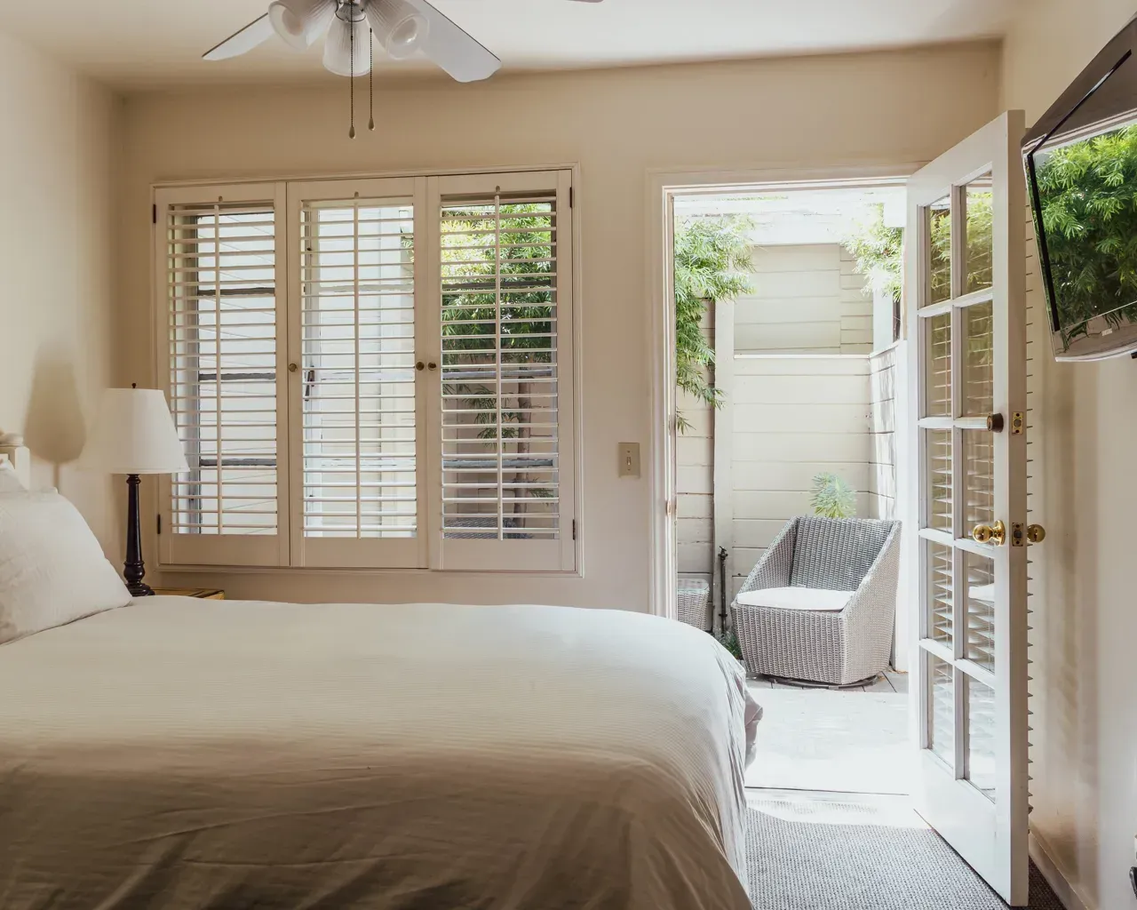 A bedroom with a white unmade bed, a table lamp, and an open glass door leading to a patio with a chair.
