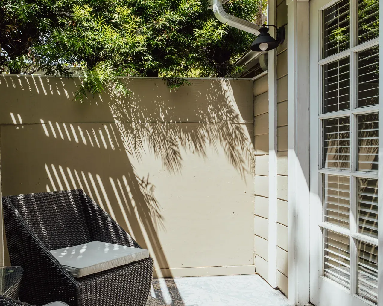 A woven chair with a white cushion sits on a patio next to a glass-paned door, partially shaded by tree leaves.