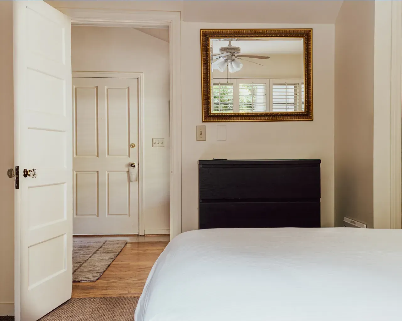 A bedroom interior featuring a white bed, a black headboard, a gold-framed mirror, and an open doorway leading to a hallway.