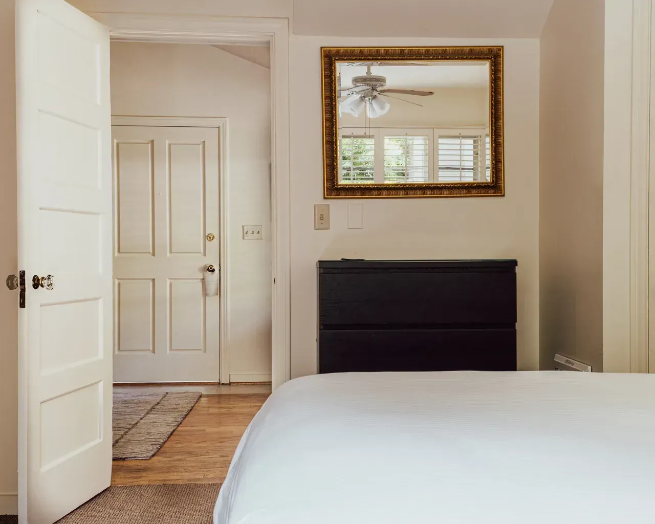 A view of a bedroom with a bed, a black headboard, and a gold-framed mirror, looking into a hallway with a white door.