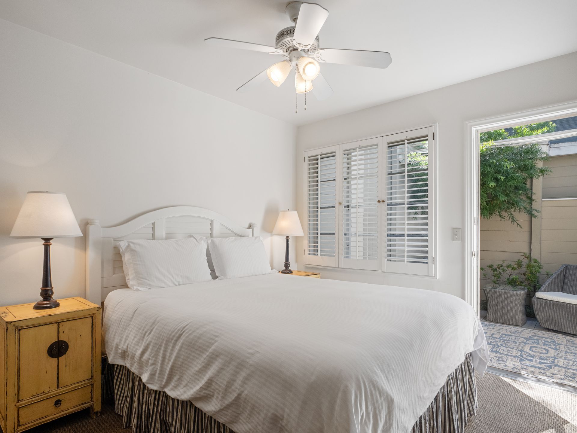A bright bedroom with a white bed, a tan bedside table with a lamp, a ceiling fan, and shutters opening to a patio.