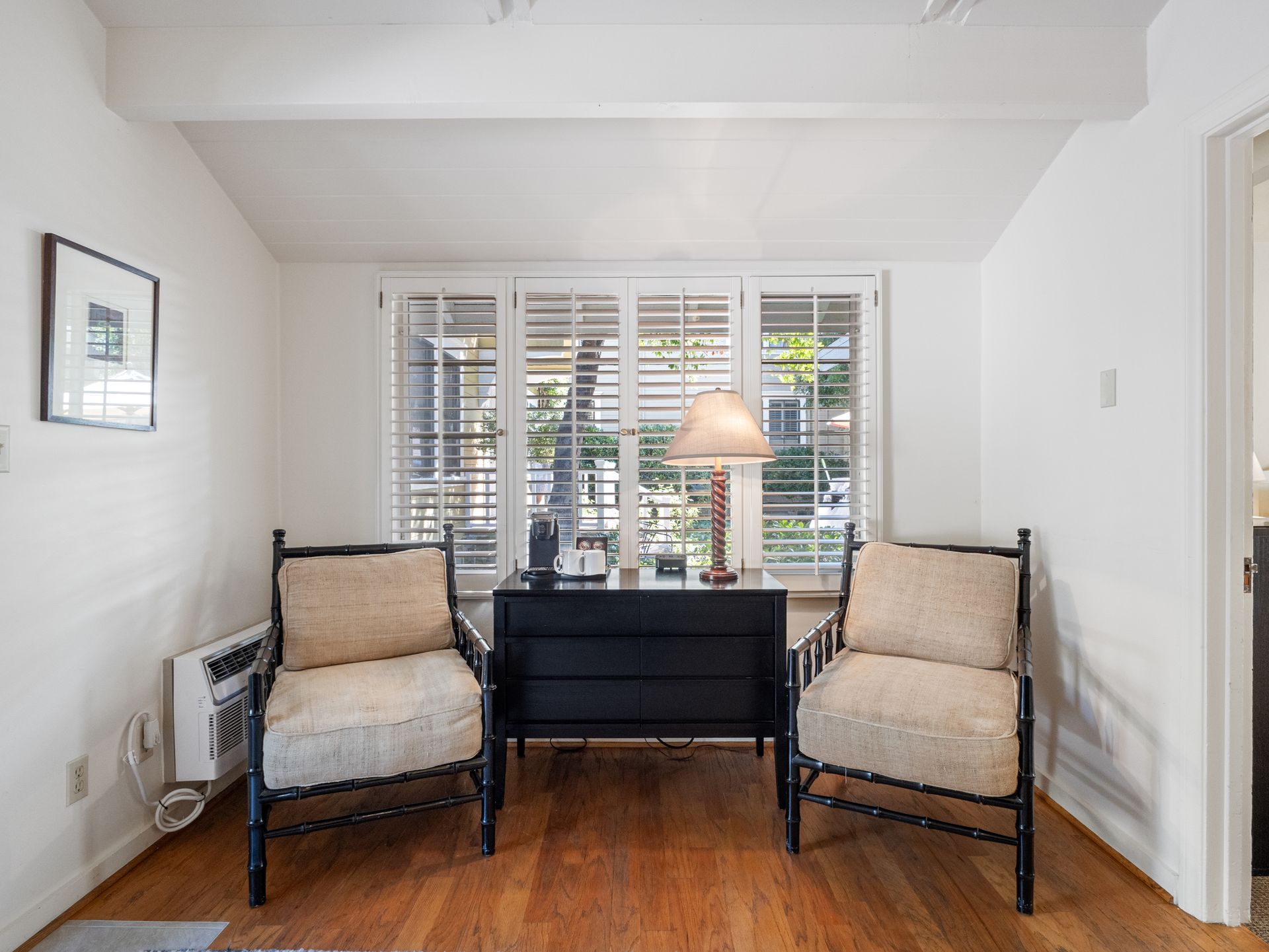 Two beige armchairs with dark frames face forward, separated by a black cabinet holding a lamp before a window.