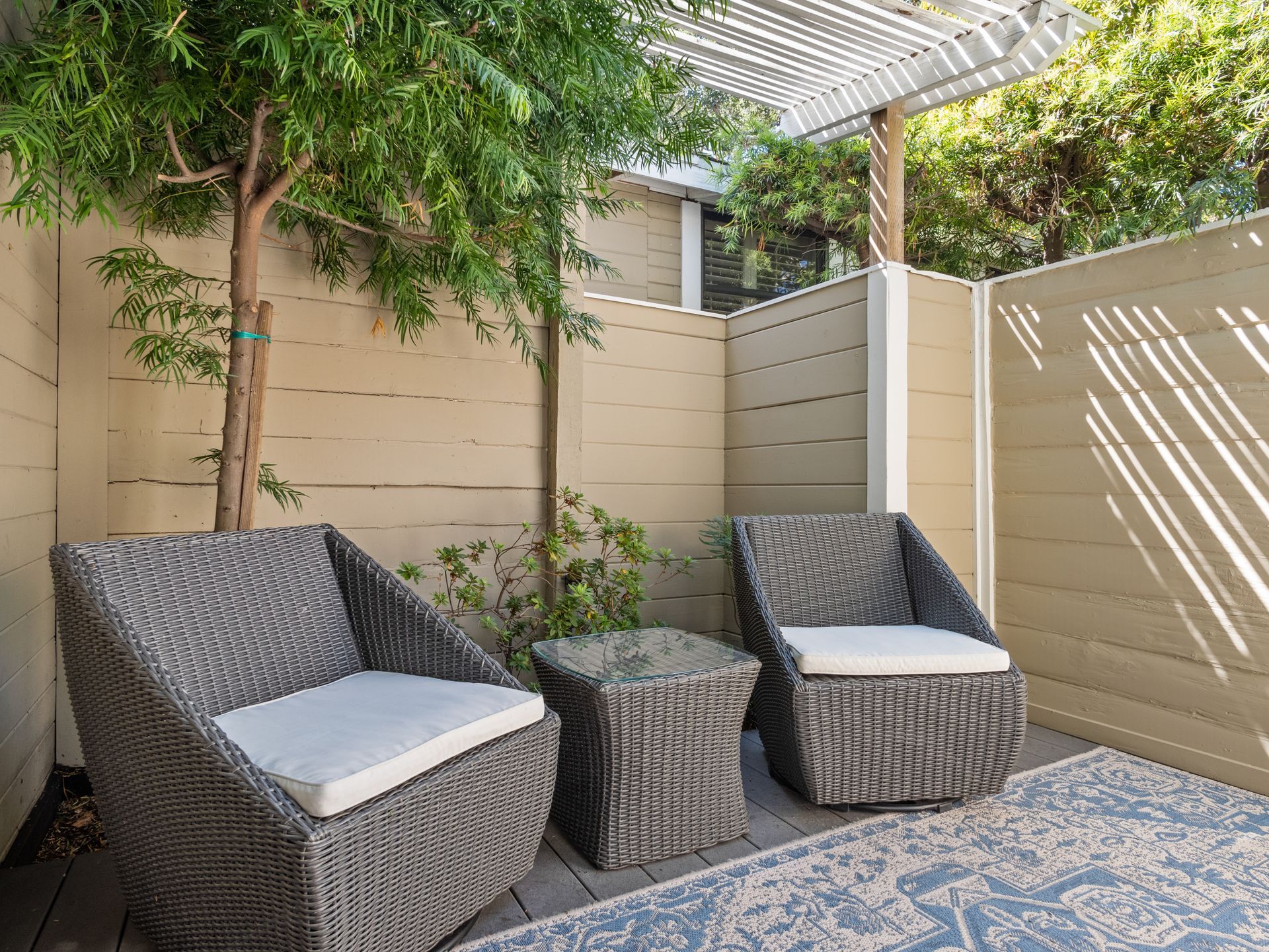 Two wicker chairs and a small table with white cushions sit on a patterned blue rug in a cozy, fenced-in patio area.