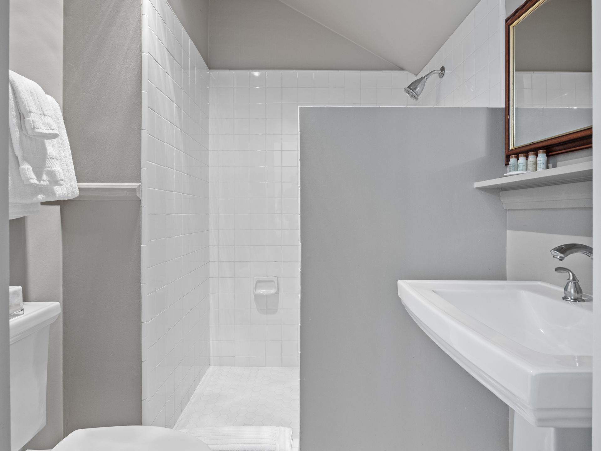 A modern bathroom with a white tiled walk-in shower, a pedestal sink, and gray walls with a mirror and hanging towels.