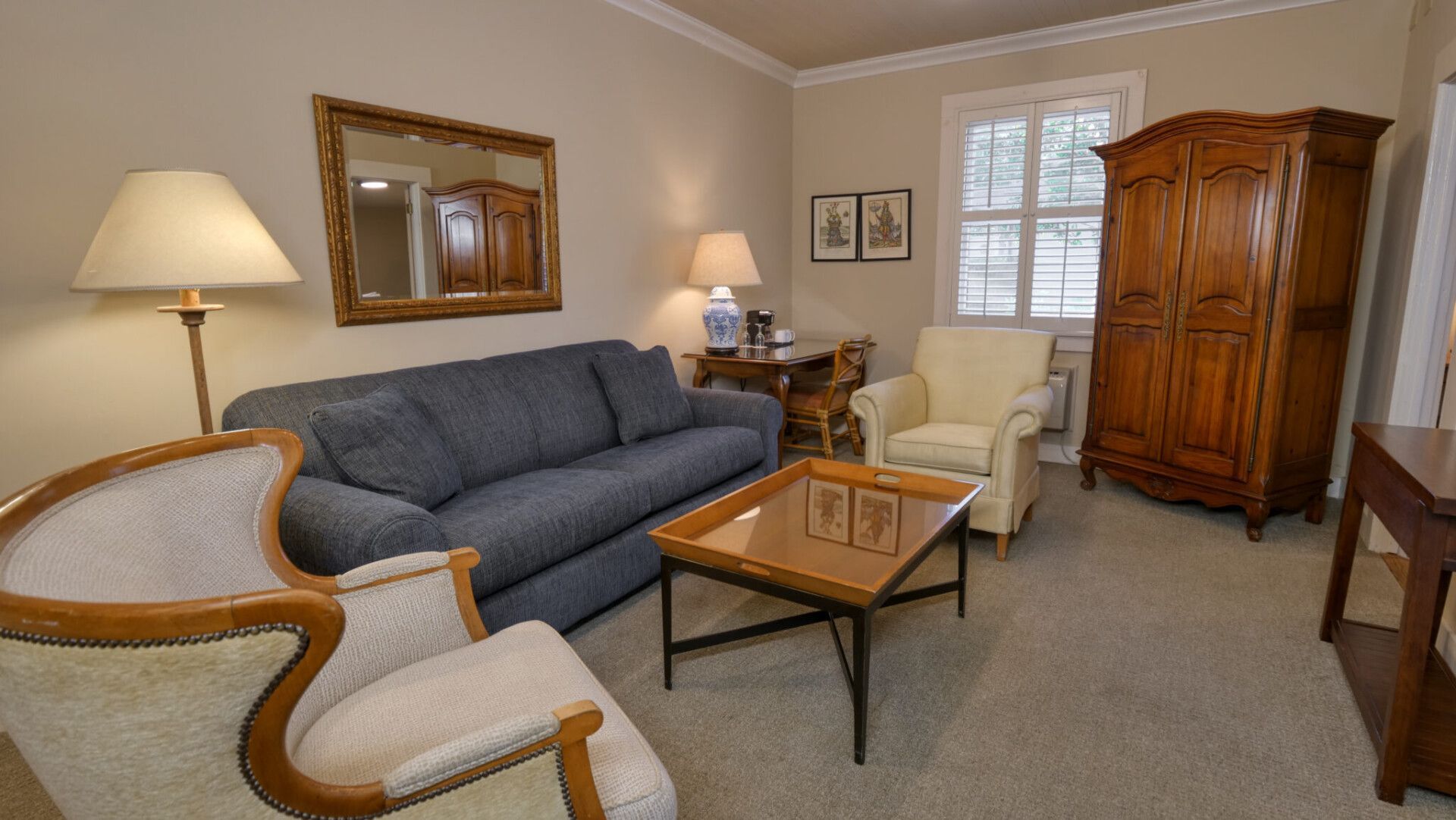 A living room with a blue sofa, light-colored armchairs, a glass coffee table, a wood armoire, and a floor lamp.