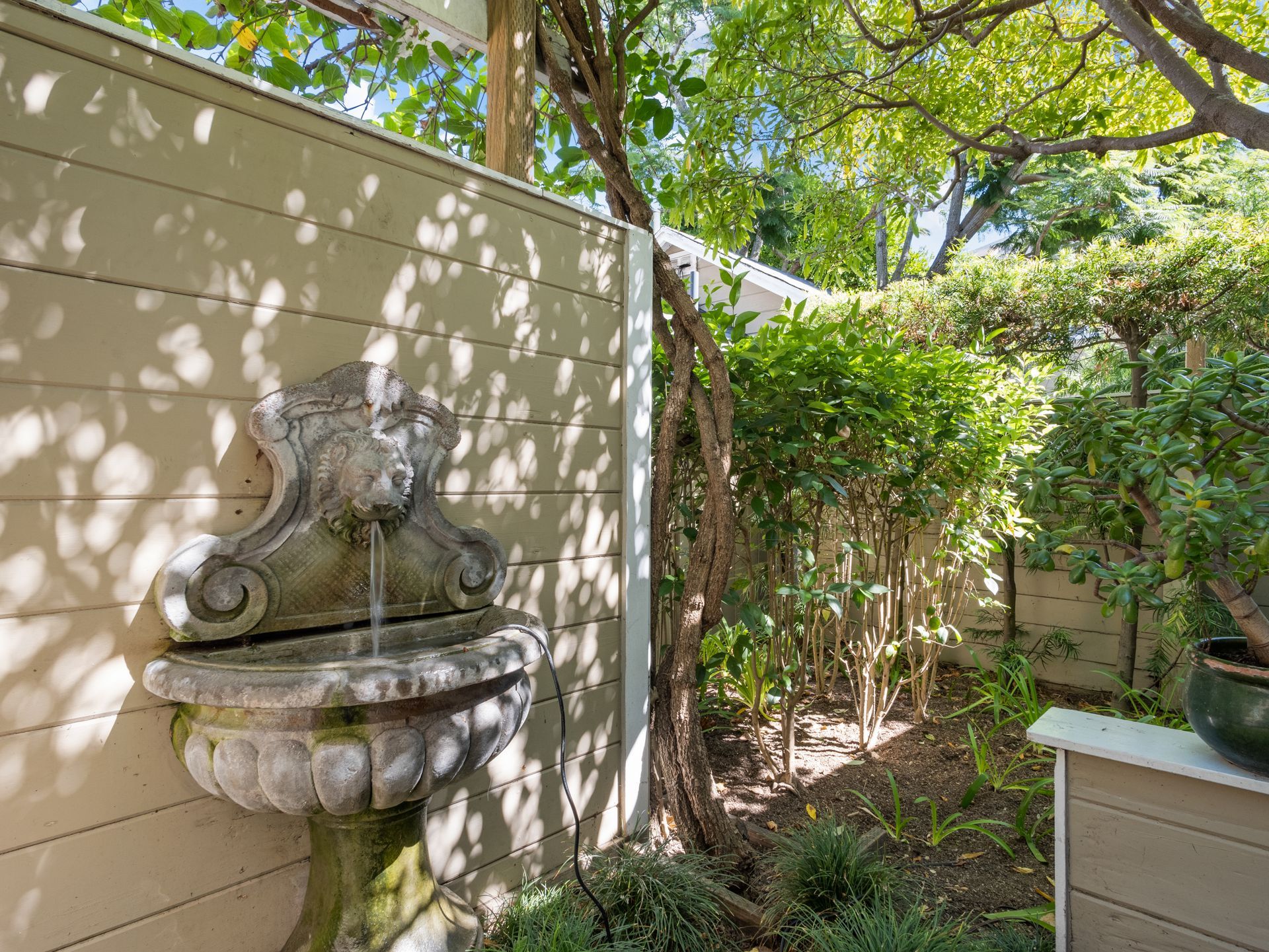 An ornate stone fountain with a lion-head spout mounted on a light-colored wooden fence in a garden setting.
