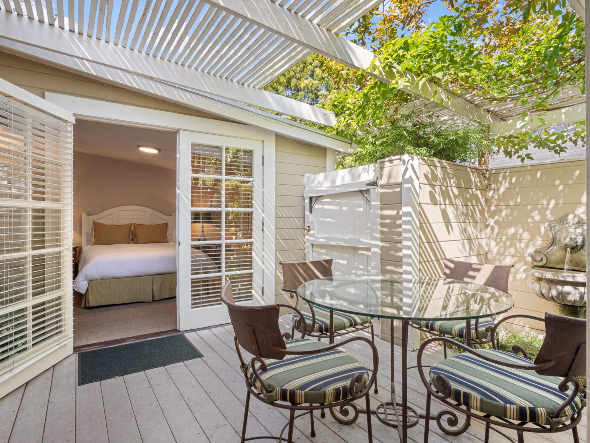 A sunlit patio with a glass-topped table and chairs leads into a bedroom through open French doors under a pergola.