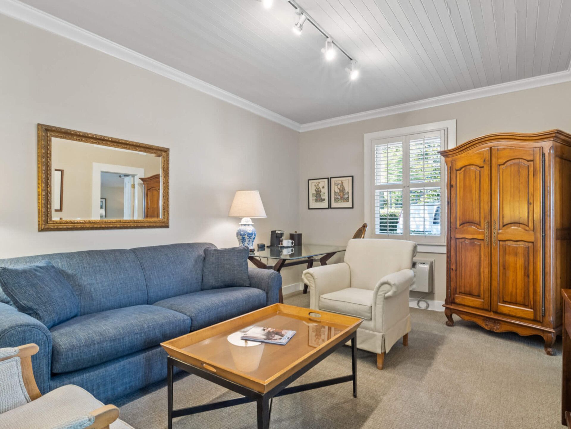 A living room with a blue sofa, white armchair, wood coffee table, gold-framed mirror, and a large wooden armoire.