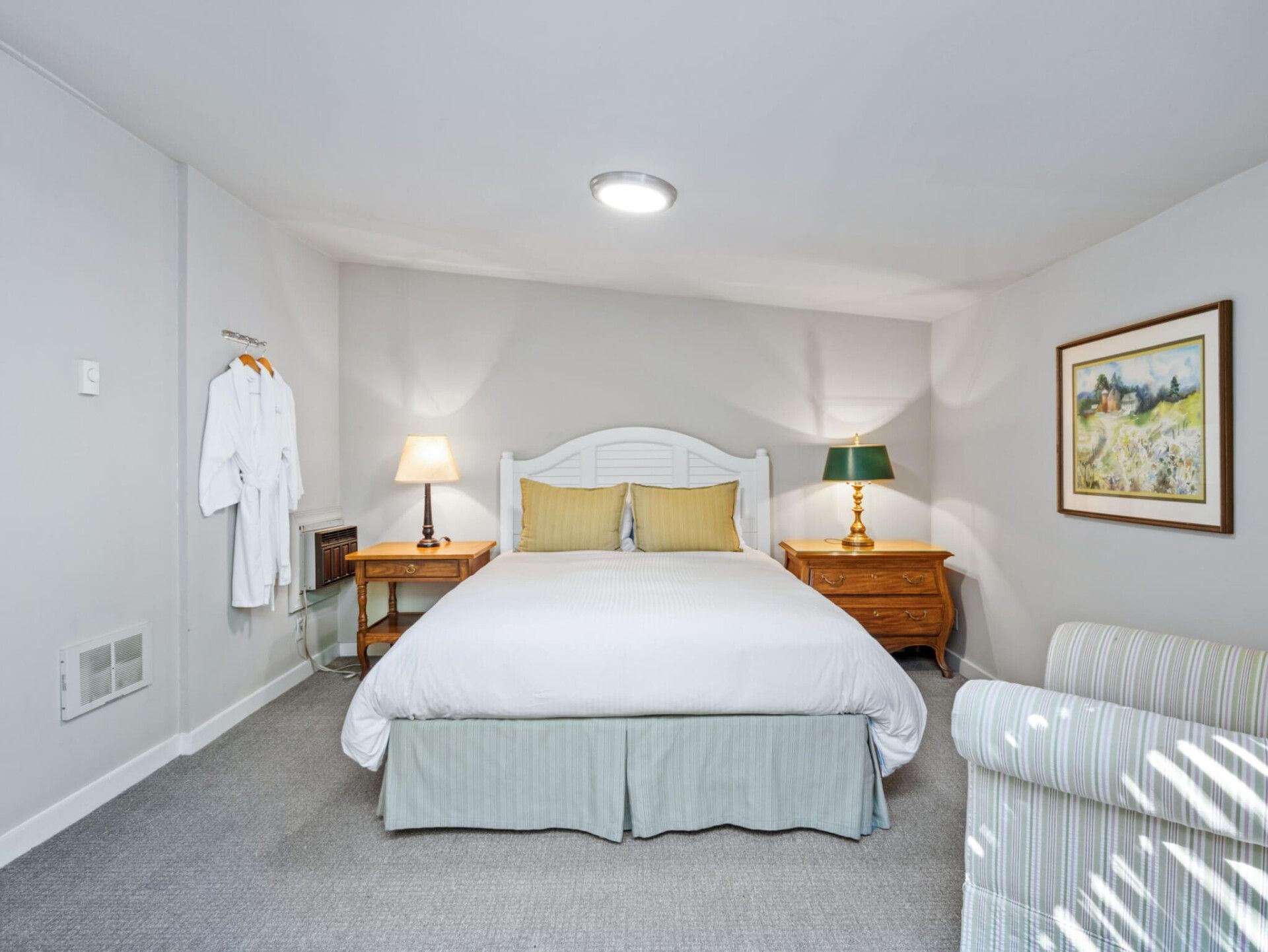 A cozy bedroom featuring a queen bed with yellow pillows, wooden nightstands, a hanging robe, and framed art.