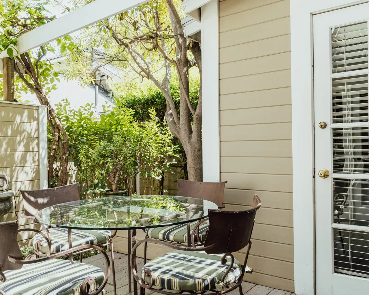 A glass-topped dining table with four chairs on a wooden patio next to a house with beige siding and a glass-paned door.