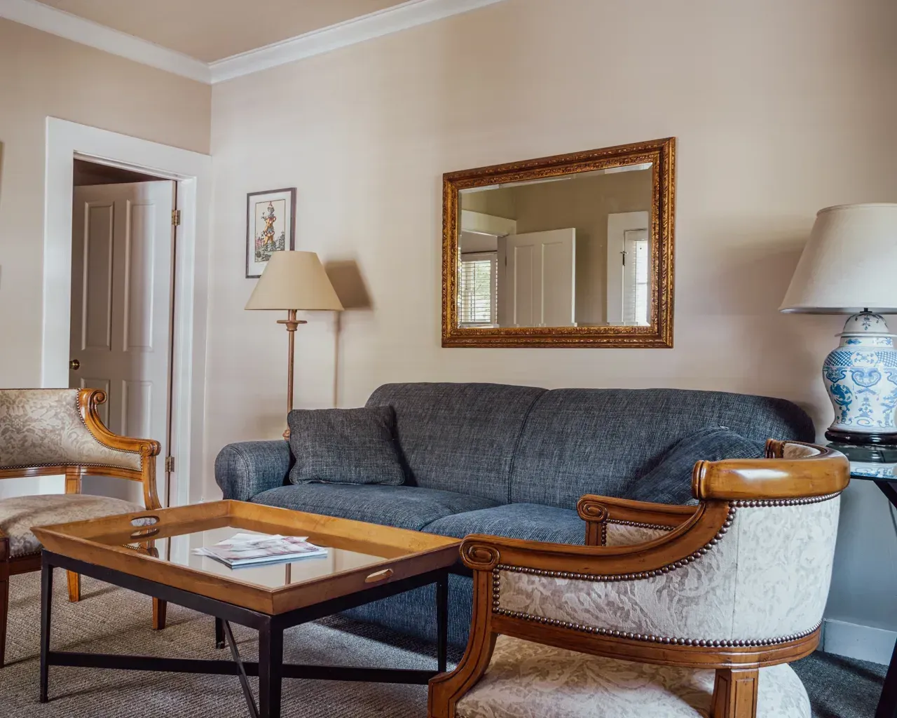 A living room with a gray sofa, a wooden coffee table, two patterned armchairs, and a gold-framed wall mirror.