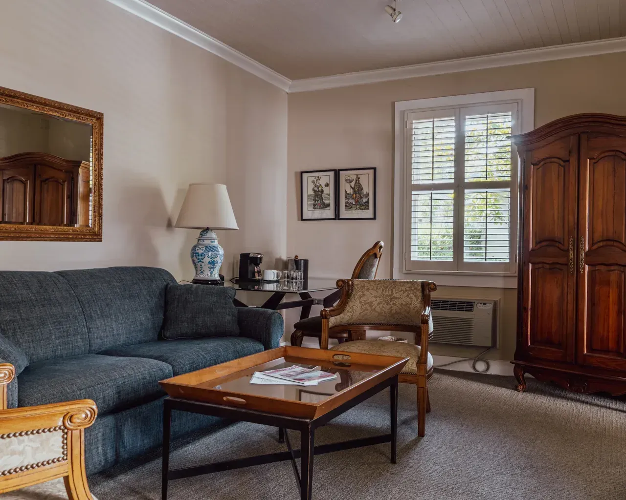 A cozy hotel room featuring a dark blue sofa, a wooden coffee table, a wooden wardrobe, and a chair near a window.