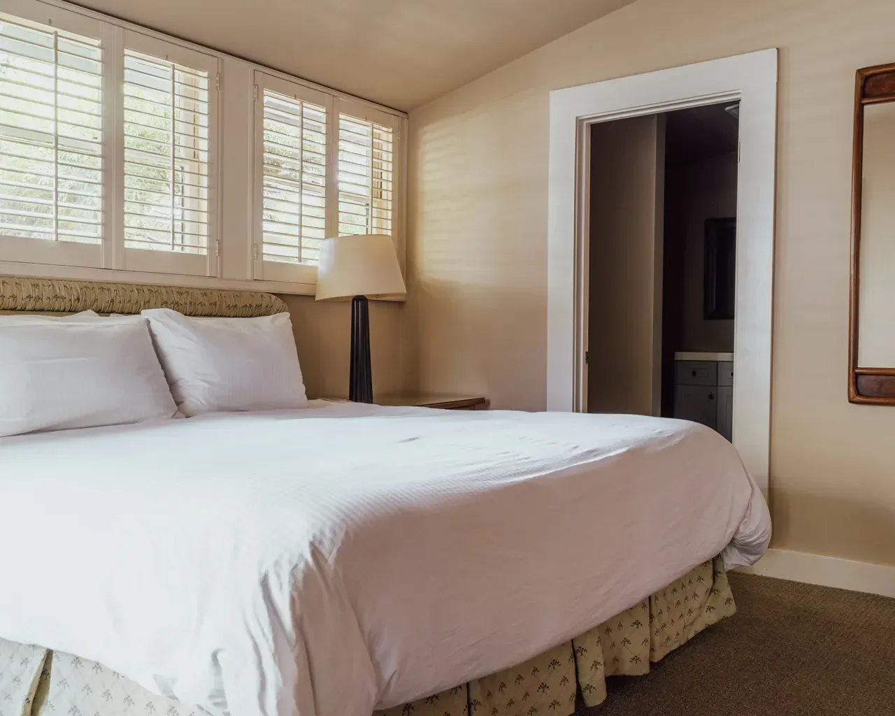 A made bed with white linens and pillows sits in a light-colored bedroom with shutters and a doorway to a bathroom.
