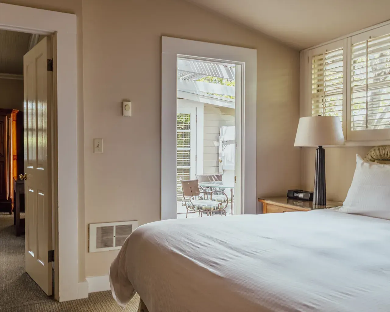 A bright bedroom with a bed, a lamp on a side table, and a door leading to a patio with outdoor furniture.