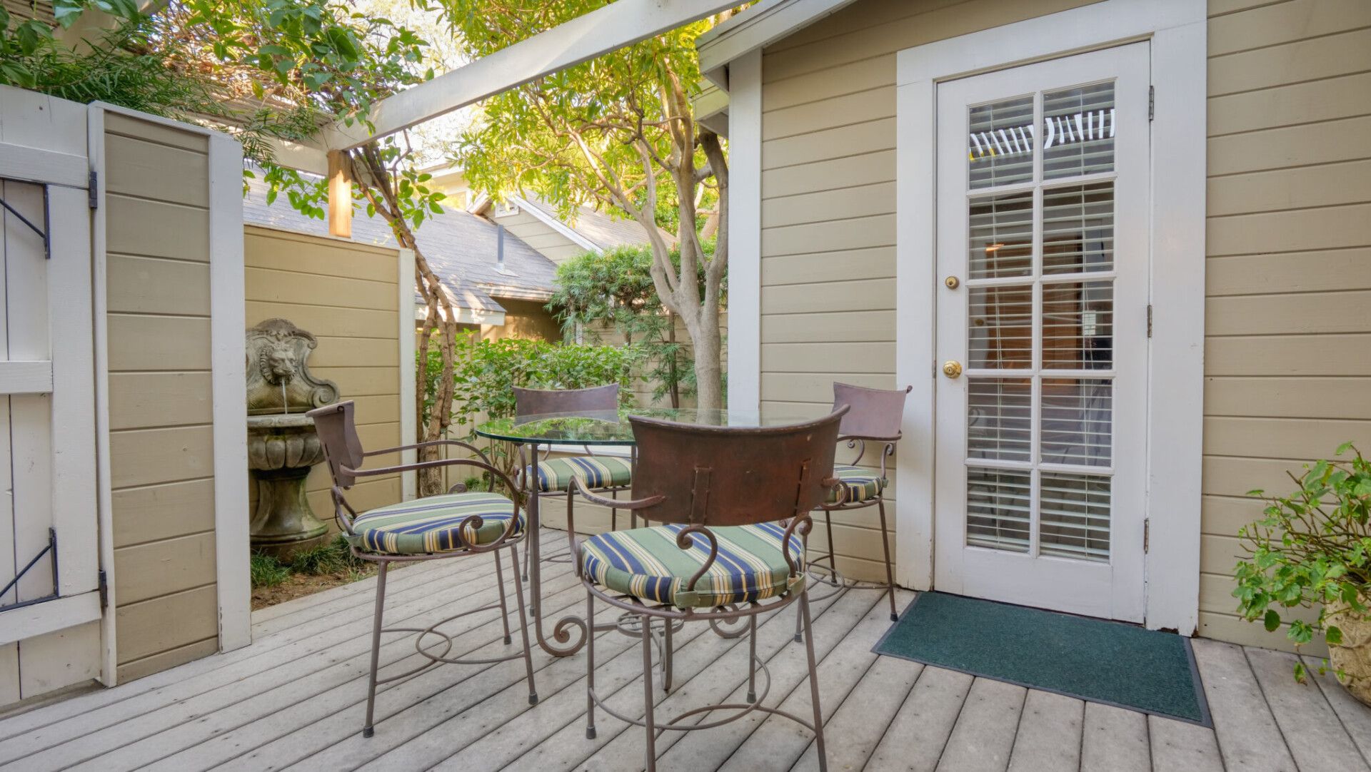 A wooden patio with a small table and chairs outside a door with white trim, set against a tan, siding-covered wall.