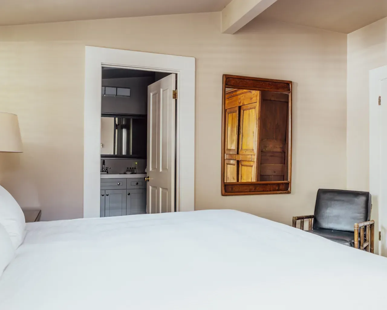 A view from a bed into a bathroom, with a dark leather armchair and a framed mirror hanging on the beige bedroom wall.