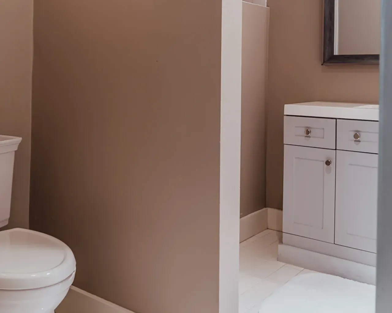 A light gray vanity with drawers sits in a bathroom with tan walls next to a white toilet.