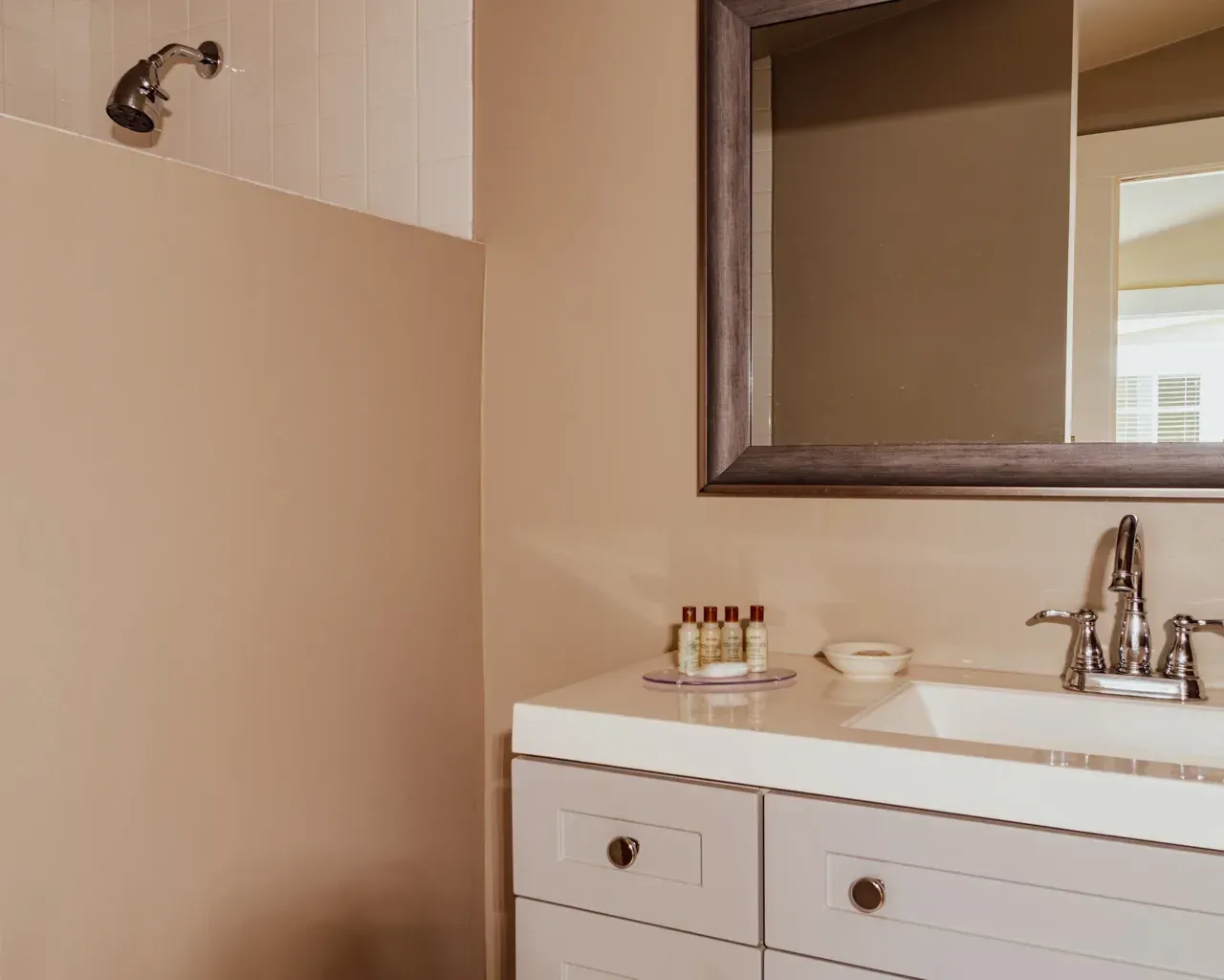 A modern bathroom vanity with a white sink, chrome faucet, small toiletries, and a framed mirror on a tan wall.
