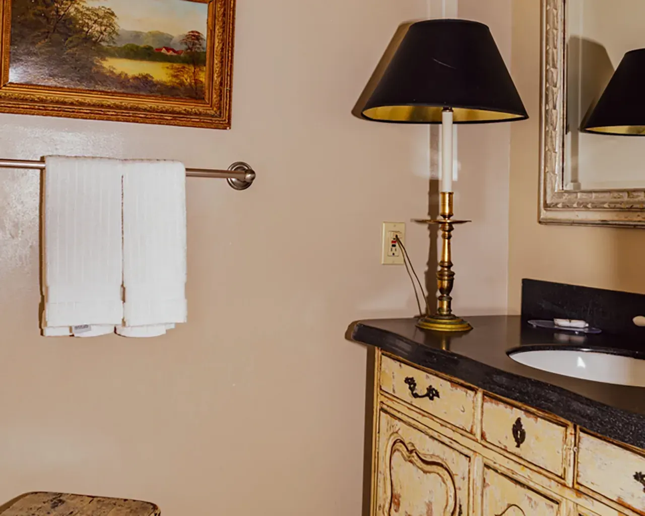 A bathroom vanity with a lamp, towels on a rack, and a framed painting on a tan wall.