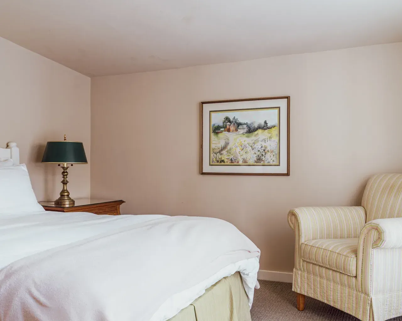 A bedroom corner with a white-covered bed, a lamp on a wooden nightstand, a textured armchair, and framed landscape art.