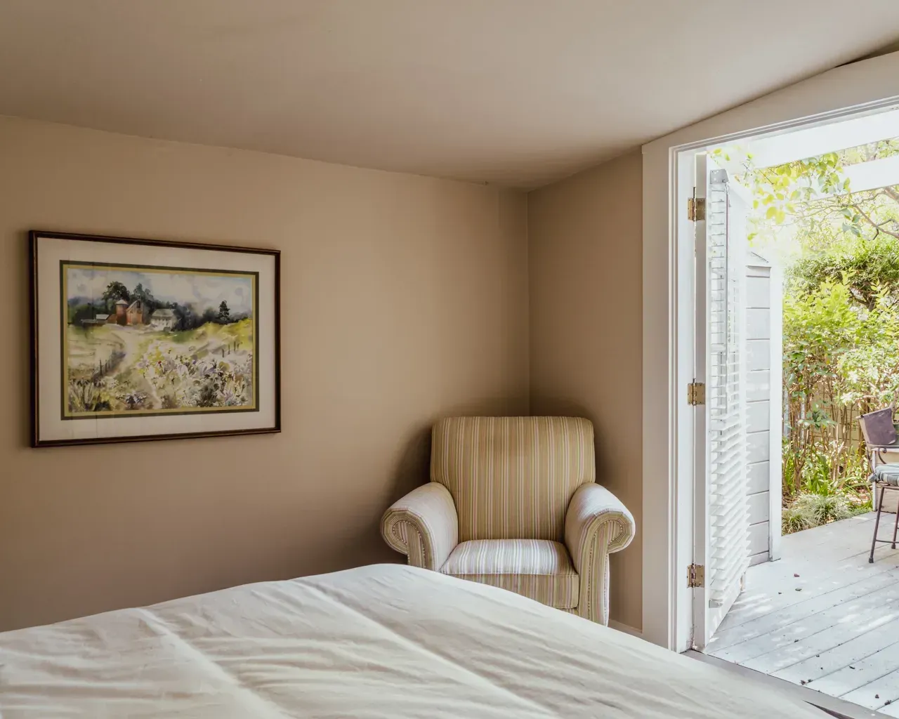 A cozy bedroom corner with a striped armchair, a framed landscape painting on the wall, and an open door to a patio.