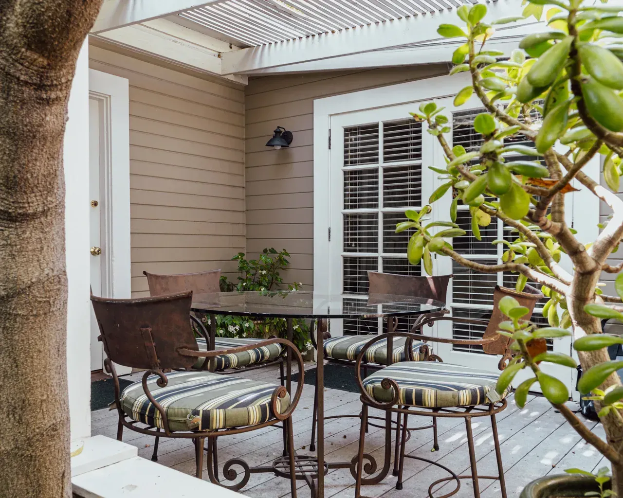 A patio with a round glass table and iron chairs, bordered by tan walls, a white door, and a vibrant green succulent plant.
