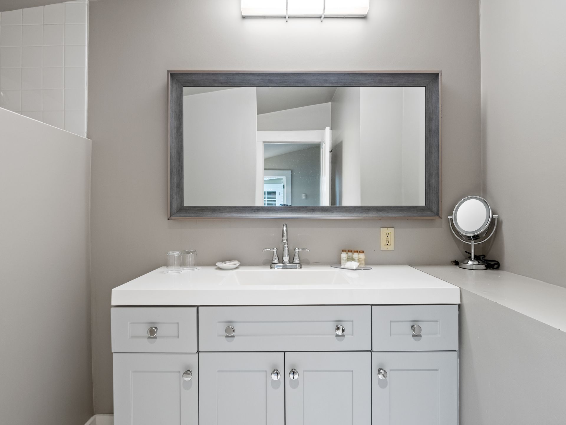 A modern bathroom vanity with a gray cabinet, a white sink, a rectangular mirror, and a small countertop makeup mirror.