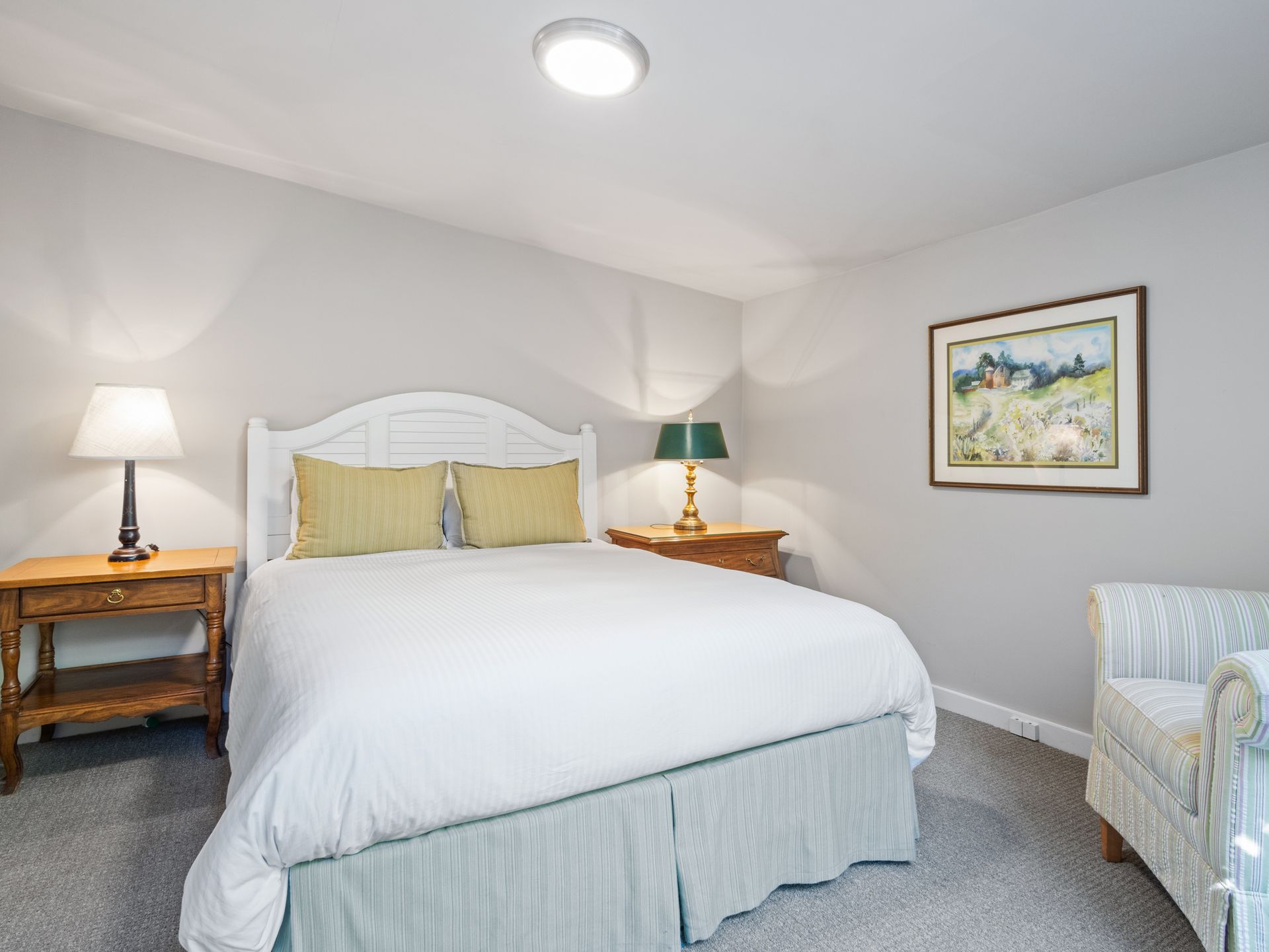 A bedroom with a queen-sized bed, white bedding, tan pillows, two wooden nightstands with lamps, and a patterned armchair.