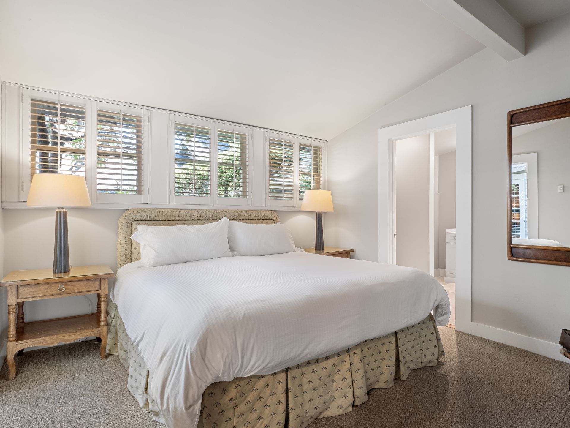 A bright bedroom with a king bed, two bedside tables with lamps, white shutters, and a wall mirror.