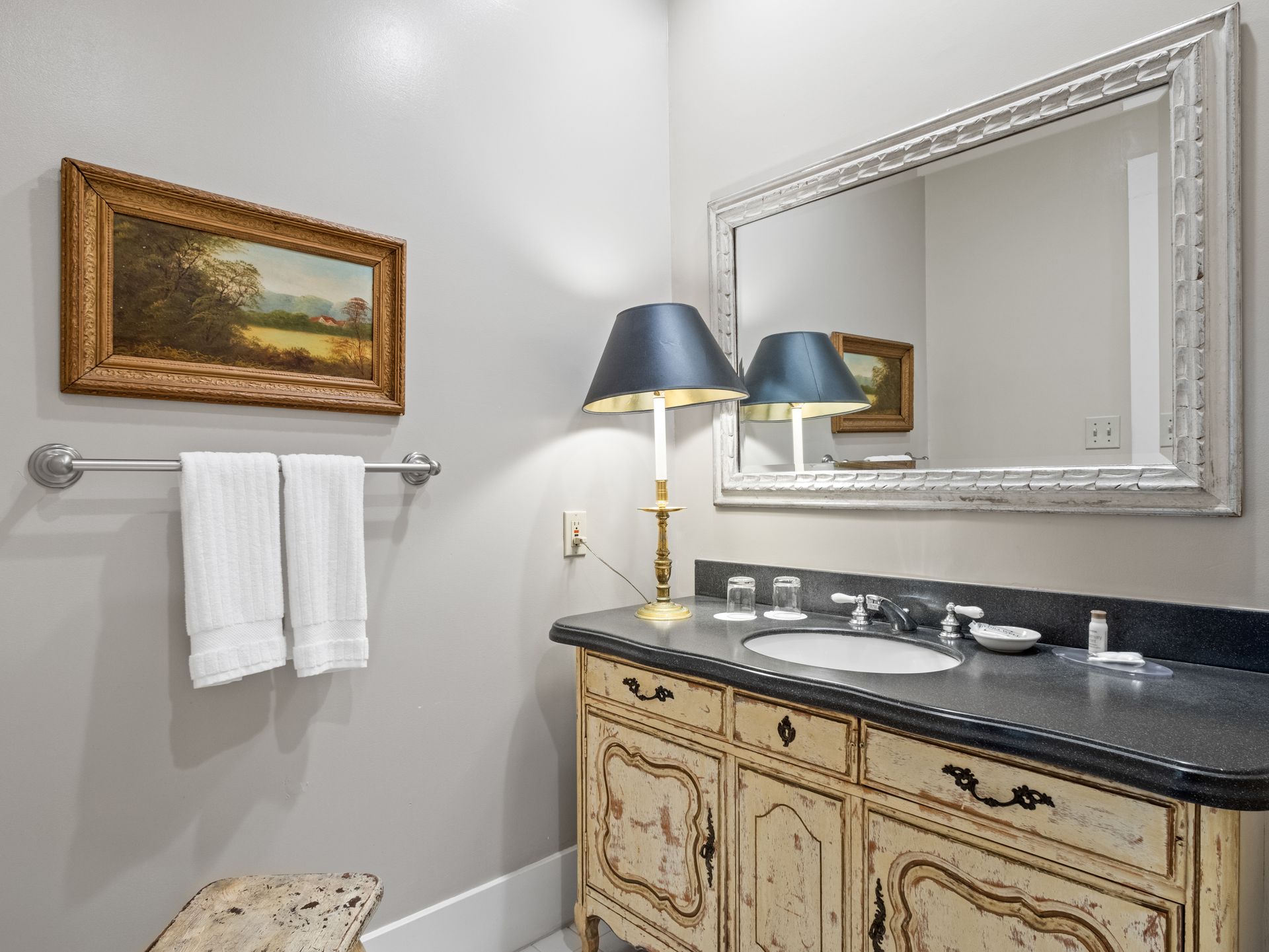 A bathroom with a white vanity, a large ornate mirror, a lamp, and two white towels hanging on a silver bar.