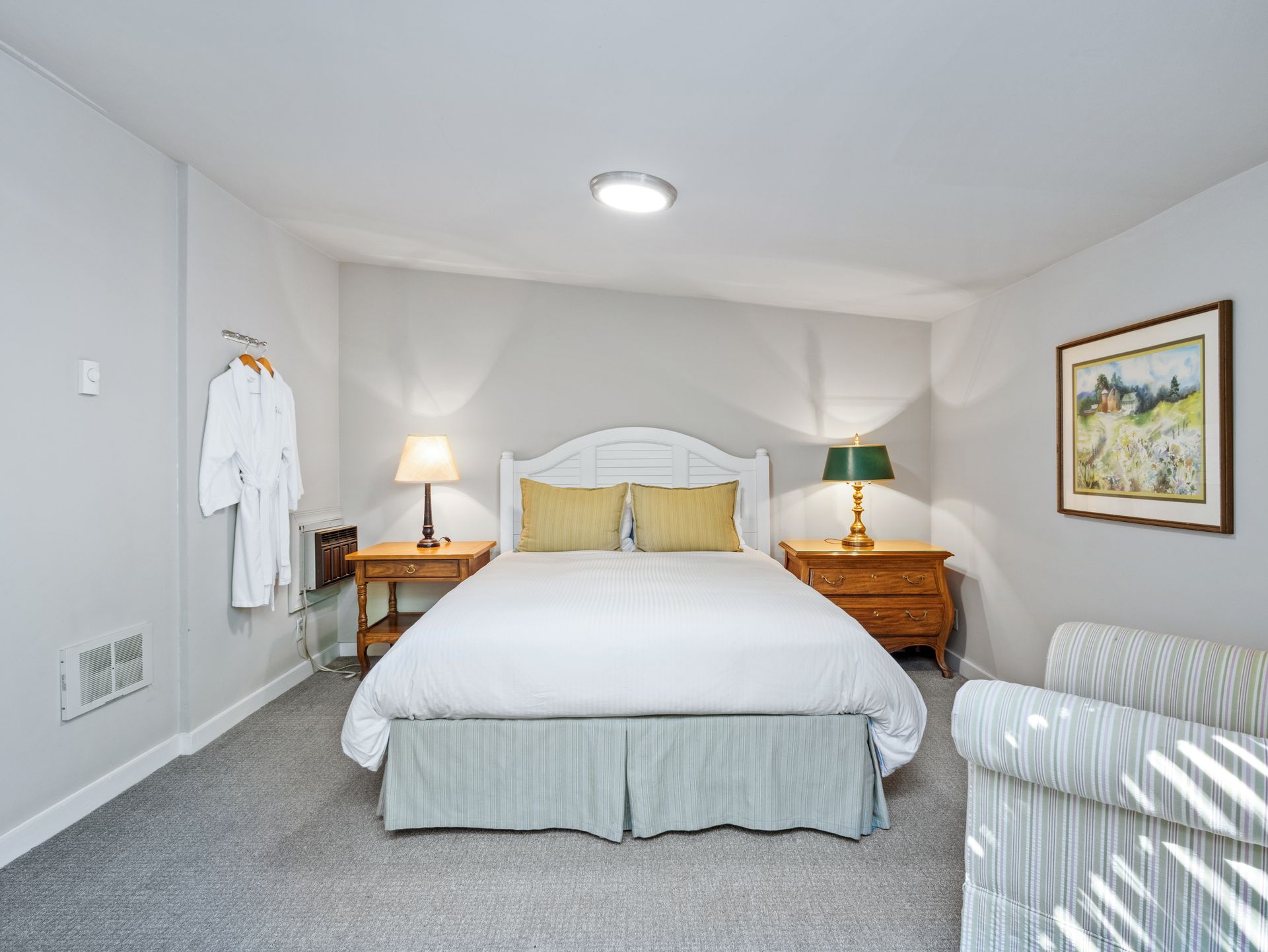A hotel room with a queen bed between two wooden nightstands, lamps, a white bathrobe on the wall, and an armchair.