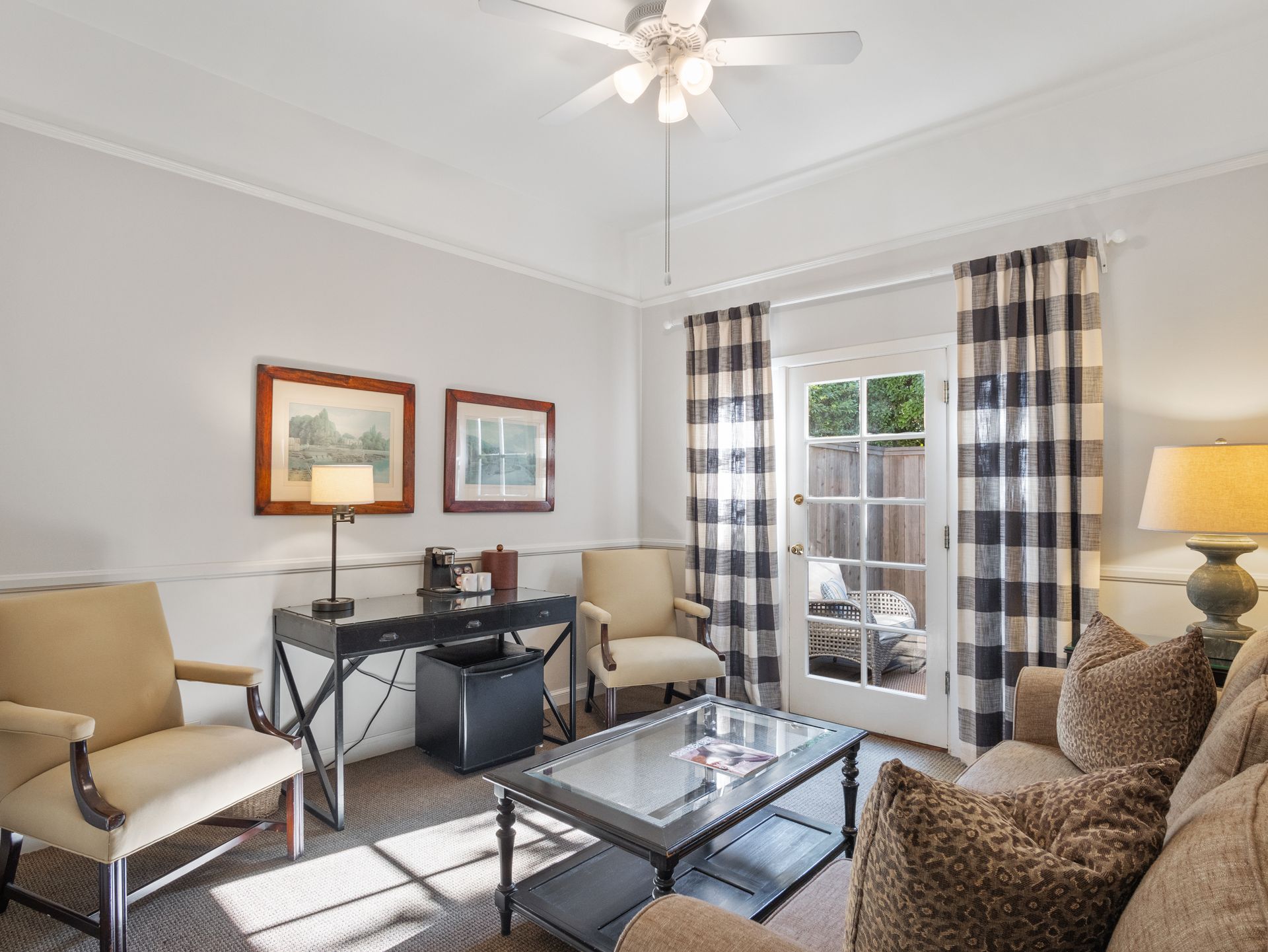 A cozy living area with beige armchairs, a glass coffee table, a desk, and buffalo check curtains by a glass patio door.