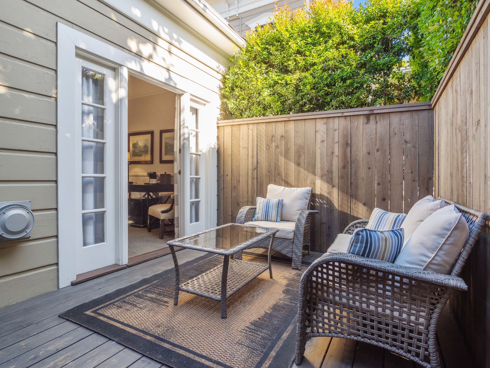 A cozy patio deck with two wicker chairs, a glass coffee table, and an outdoor rug next to a light-colored home exterior.