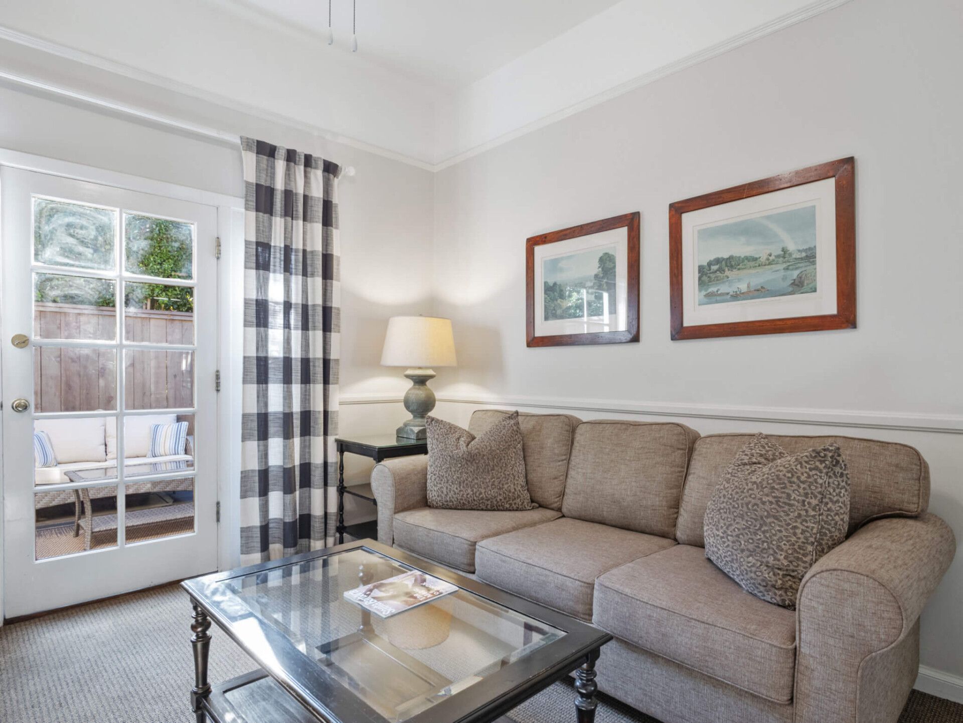 A living room with a beige sofa, glass coffee table, lamp, and framed wall art next to a door with plaid curtains.