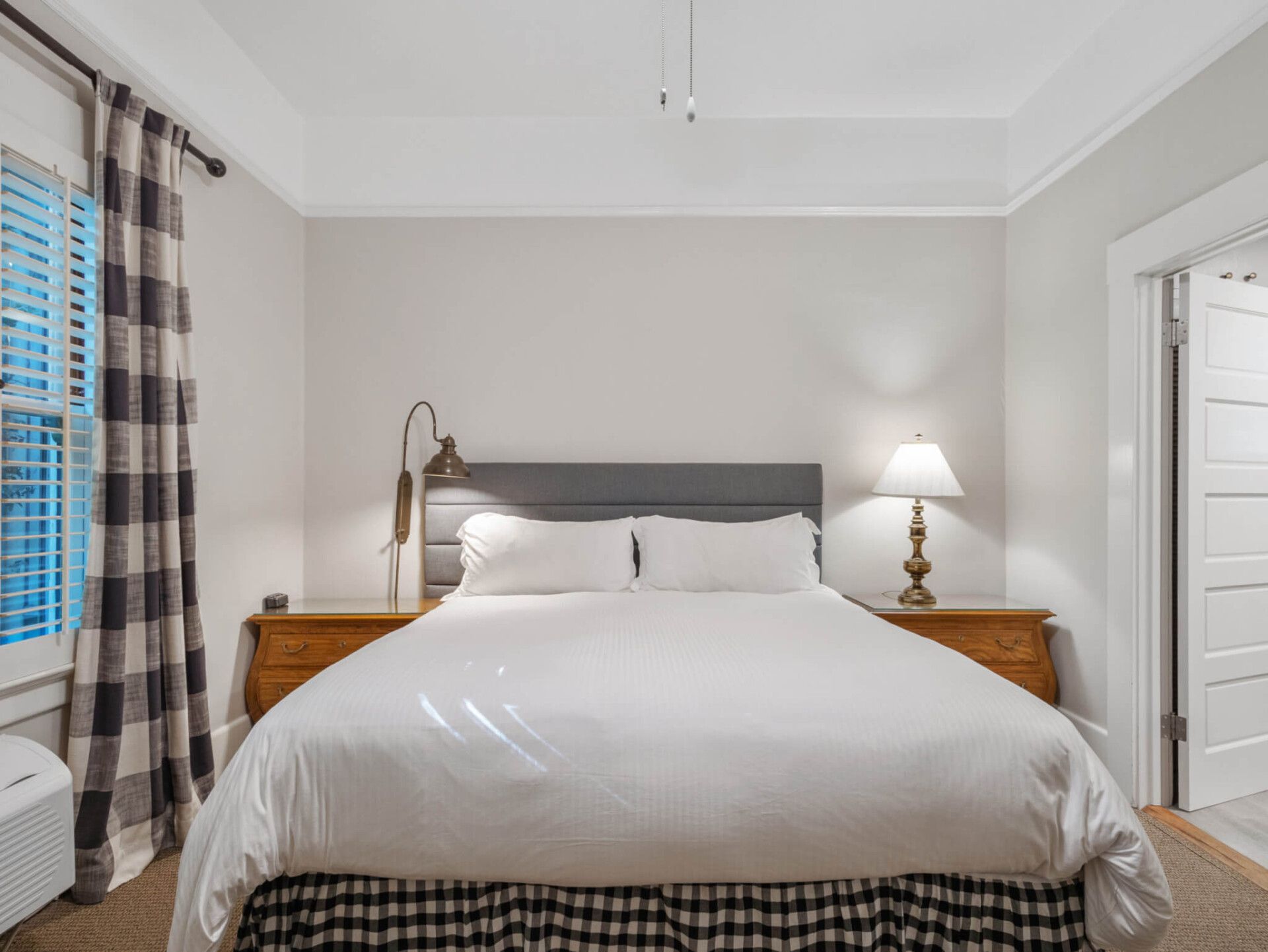 A bedroom with a queen bed featuring a grey headboard, white bedding, a buffalo check bed skirt, and two wooden nightstands.