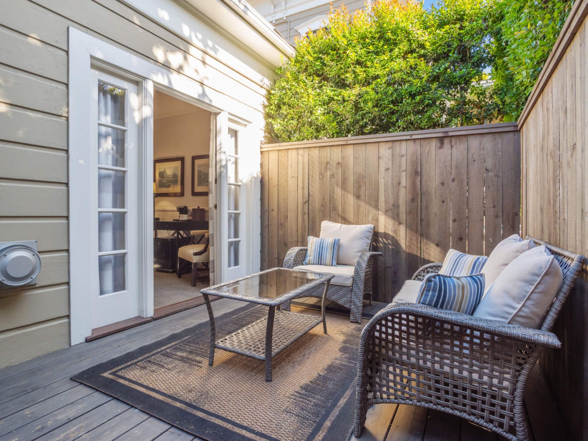 A patio with a wicker loveseat, armchairs, and glass coffee table on a deck, with double doors leading into a room.