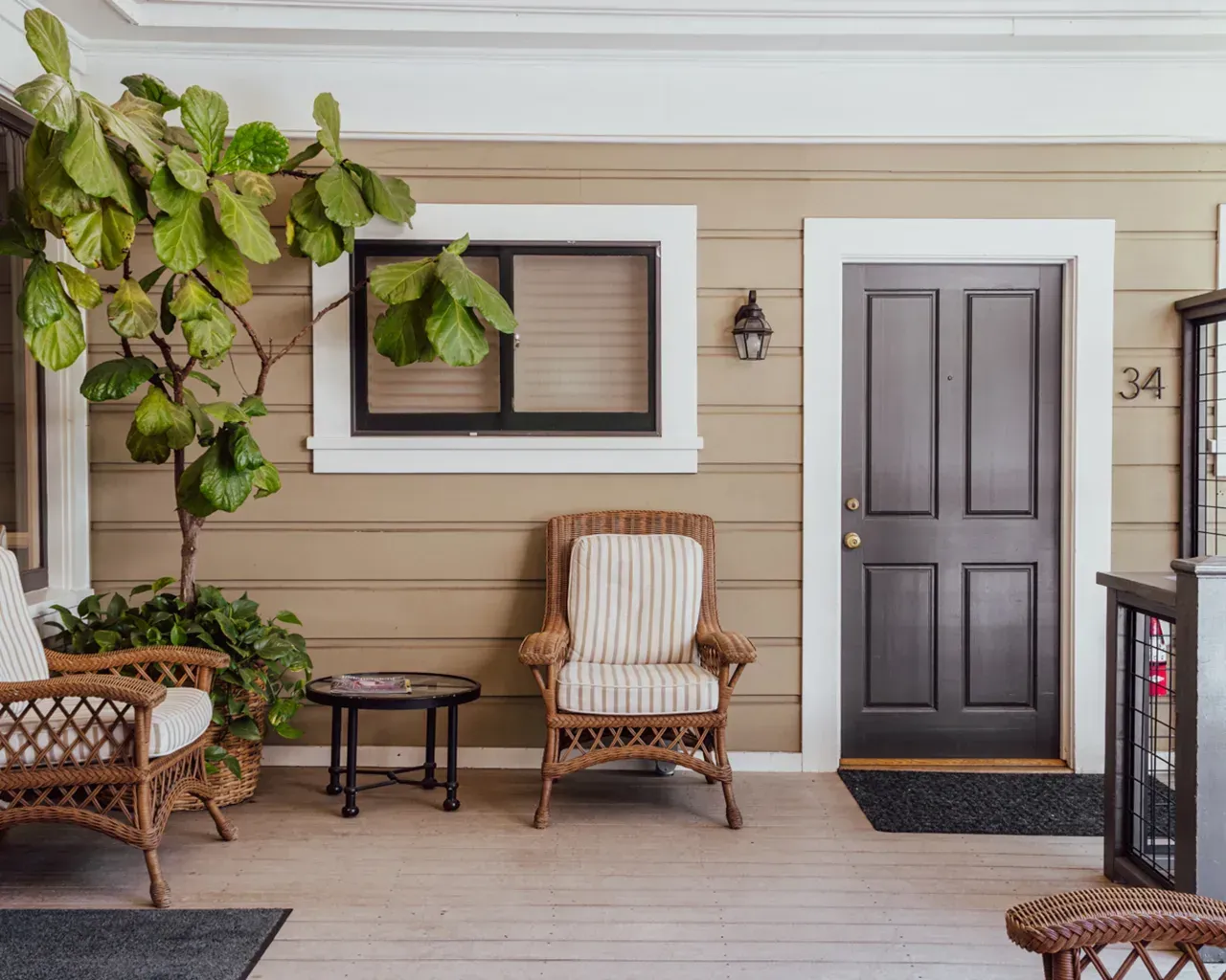 A covered front porch with two wicker chairs, a small table, a fiddle-leaf fig plant, and a dark brown door numbered 34.