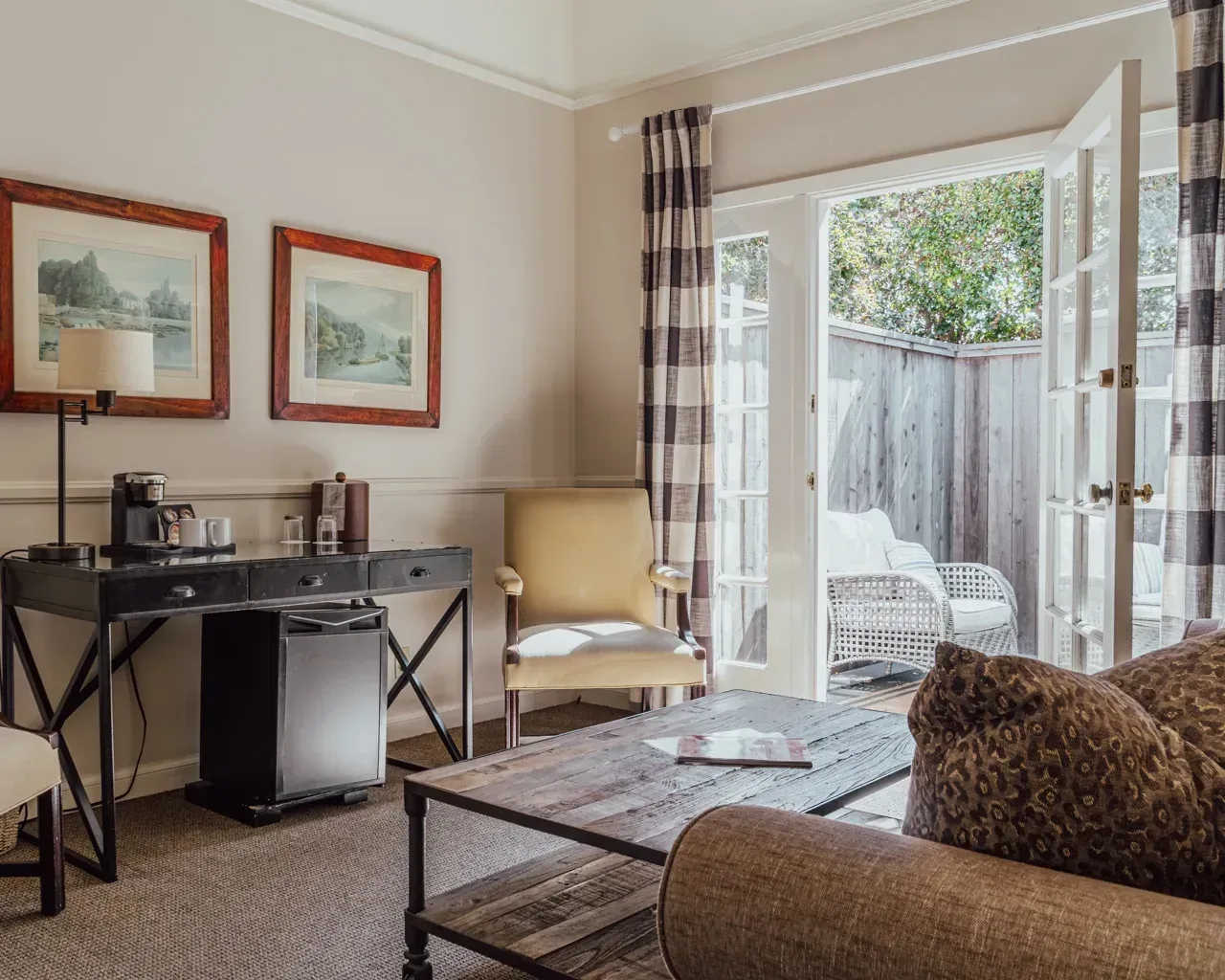 A cozy hotel room featuring a dark desk, yellow armchair, wooden coffee table, and an open glass door leading to a patio.