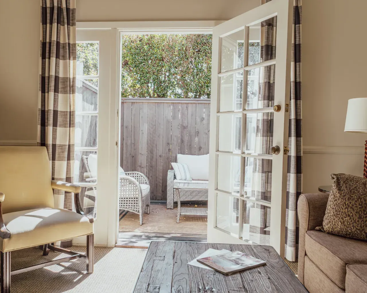 A cozy room with a yellow chair, a couch, and open French doors leading to a patio with wicker furniture.
