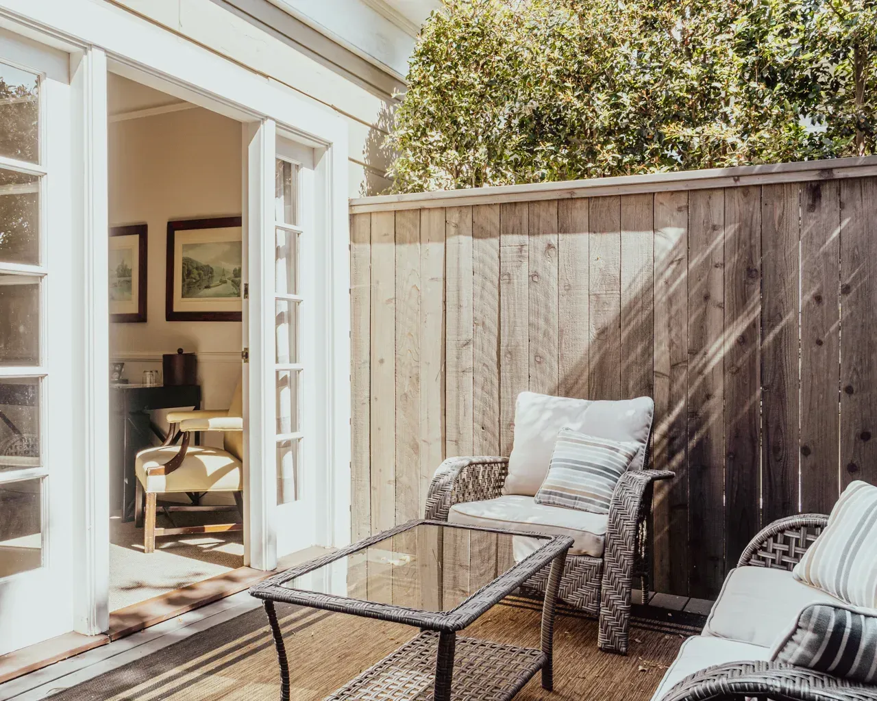 Wicker patio furniture with cushions sits on a deck outside an open glass door, beside a tall wooden privacy fence.