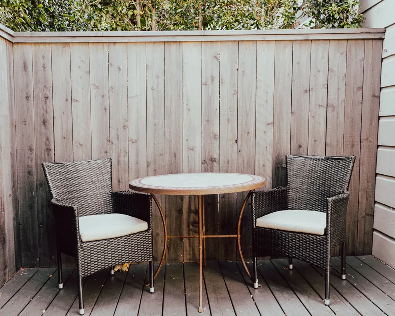 Two dark wicker chairs with light cushions and a small matching bistro table set on a deck against a tall wooden fence.