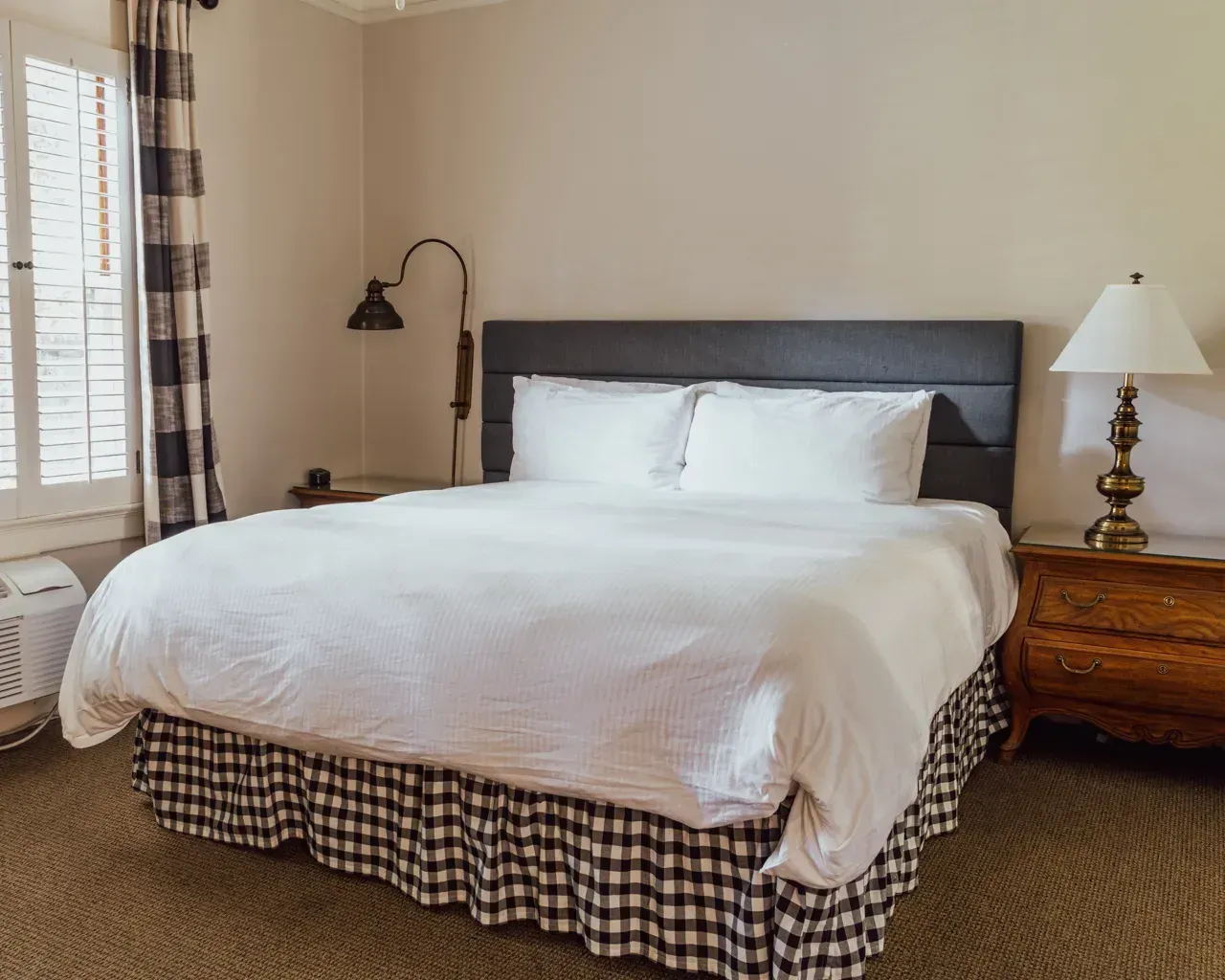 A made bed with white bedding and a black-and-white buffalo check bed skirt in a room with a lamp and side table.