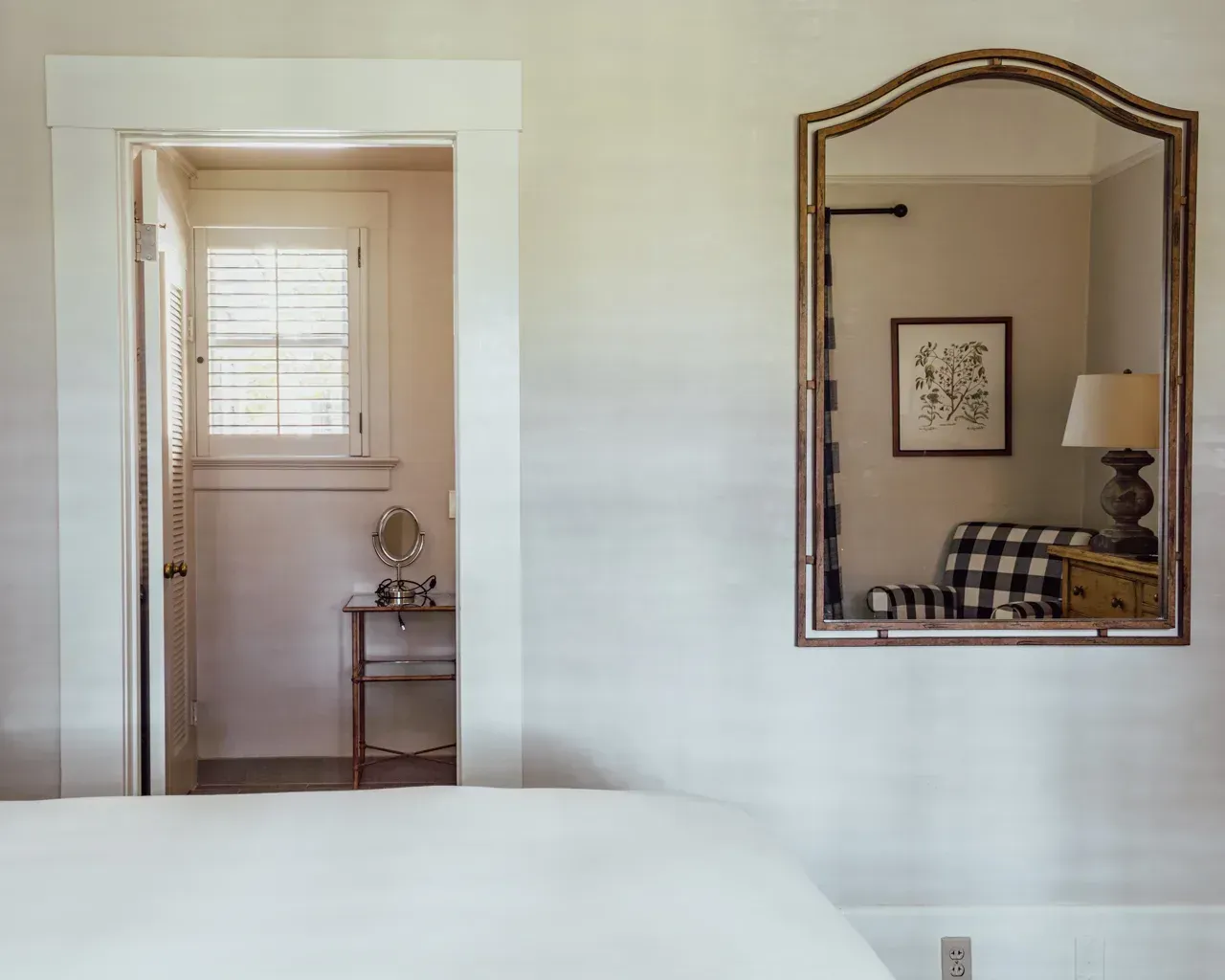 A bedroom scene featuring a white-framed doorway to a bright room and a decorative arch-top wall mirror reflecting a chair.