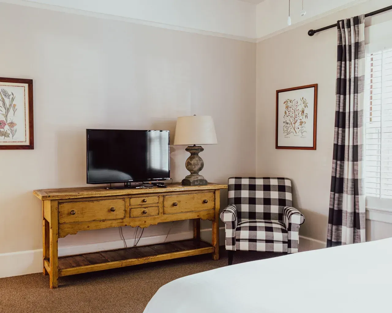 A rustic wooden dresser with a TV and lamp stands beside a black and white checkered armchair in a neutrally toned room.